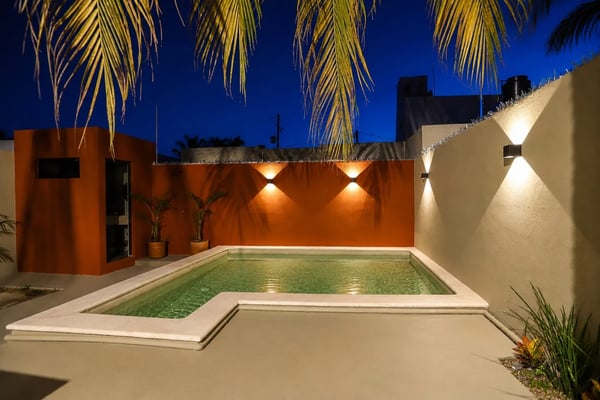 Rooftop courtyard at dusk with illuminated pool, orange and cream walls, palm fronds overhead, and city skyline beyond.