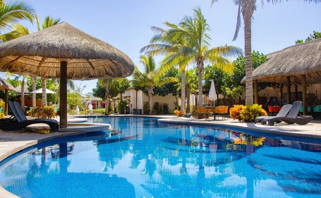 Modern beachfront villa with turquoise pool, wooden deck, thatched-roof pergola, and white building with dark railings
